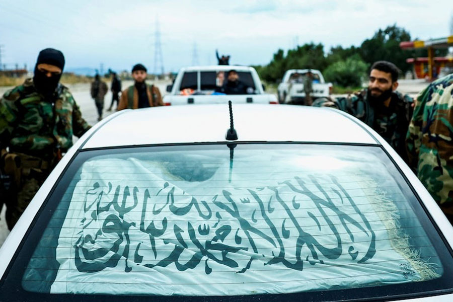 Syrian security forces stand alongside a vehicle on on the Latakia-Jablah highway in western Syria on March 21, 2025. The flag in the vehicle reads “There is No God but Allah. Muhammad is God’s Messenger”. A Reuters investigation found that thousands of forces carried out a wave of deadly attacks on Alawite villages in the region, leaving nearly 1,500 dead from March 7-9, 2025.