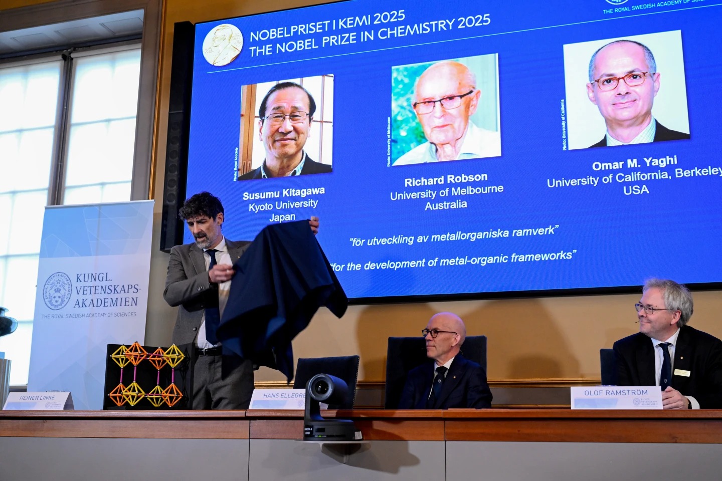 Chair of the Nobel Committee for Chemistry Heiner Linke makes a demonstration, next to Secretary General of the Swedish Academy of Sciences Hans Ellegren, and Member of the Nobel Committee for Chemistry Olof Ramstrom, right, after they announce Susumu Kitagawa, Richard Robson and Omar Yaghi, on screen behind, as the recipients the Nobel Prize in Chemistry, at the Nobel Assembly of the Karolinska Institutet, in Stockholm, Sweden, Wednesday, Oct. 8, 2025. (Fredrik Sandberg/TT News Agency via AP)