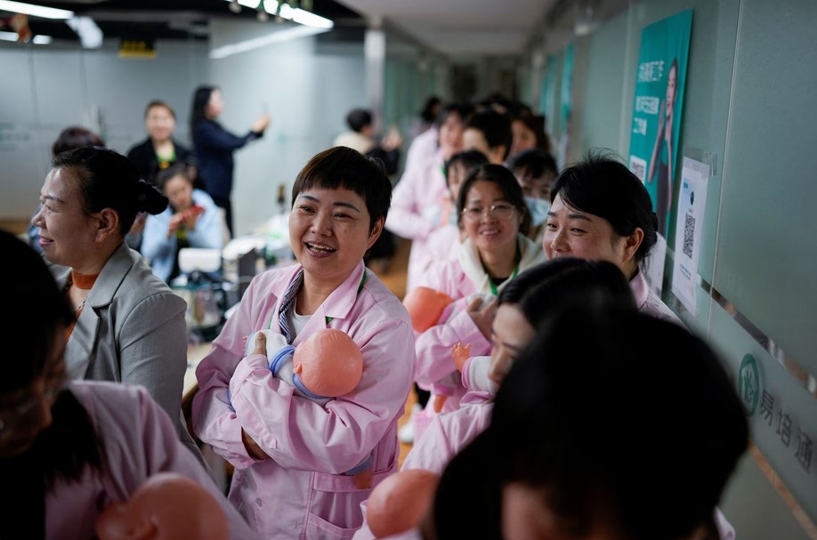 Women holding plastic baby dolls, wait to enter a classroom before a nursing skills class for confinement carers, at Yipeitong training centre in Shanghai, China March 2, 2023. REUTERS/Aly Song