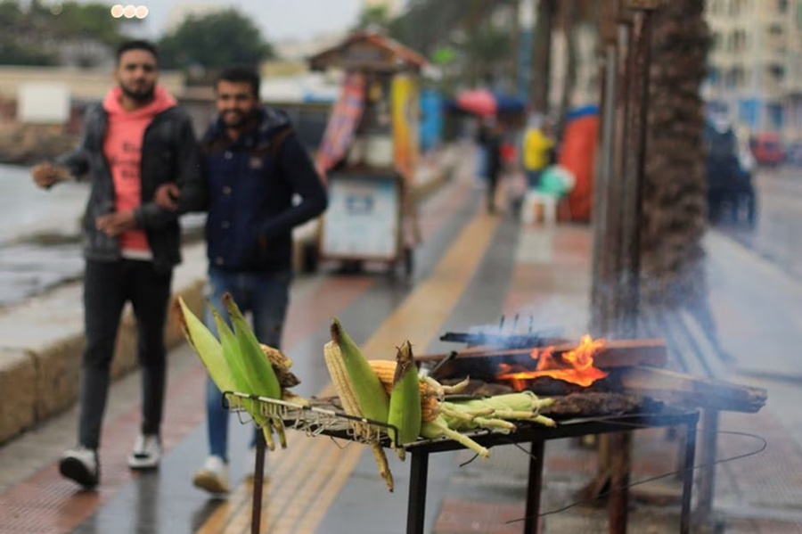 People walk next to a corn seller during rainy weather in the Mediterranean port city of Alexandria, Egypt on December 31, 2021 — Reuters